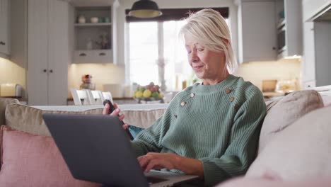 happy senior caucasian woman sitting on sofa in living room, using laptop and smartphone