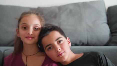 kid brother rests head on sister shoulder playfully as she smiles warmly both looking directly at camera seated indoors against soft grey sofa showing sweet sibling bond