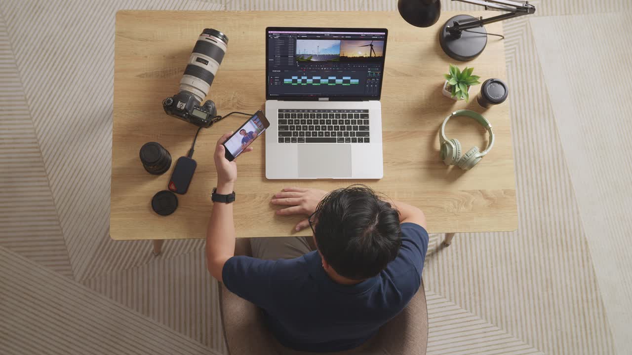 vista superior de un editor masculino agitando la mano y teniendo una llamada de video en el teléfono inteligente mientras está sentado en el espacio de trabajo usando una computadora portátil al lado de la cámara editando el video en casa