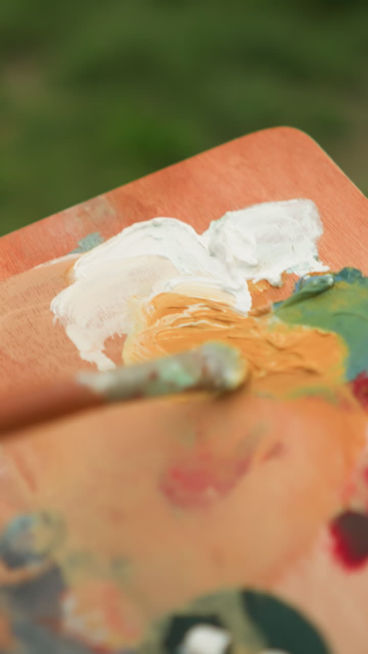 A close-up shot of an artist's hand holding a palette and mixing paint with a small brush. The vibrant colors blend together as the artist prepares to create