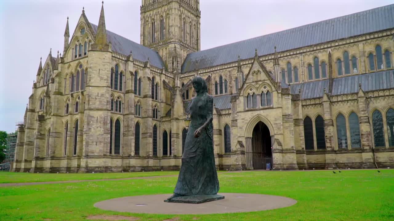 Wide shot of Salisbury Cathedral with a bronze statue of a woman in the foreground, capturing gothic architecture and open green space on a cloudy day.
