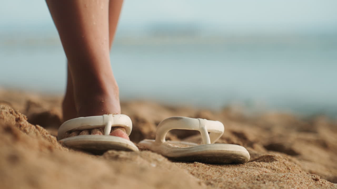 Infradito bianche sulla sabbia in primo piano, persona che avanza lasciando impronte fresche, granelli di sabbia si attaccano alle dita e alle piante dei piedi, oceano sfocato e orizzonte morbido sullo sfondo, la calda luce di mezzogiorno trasmette