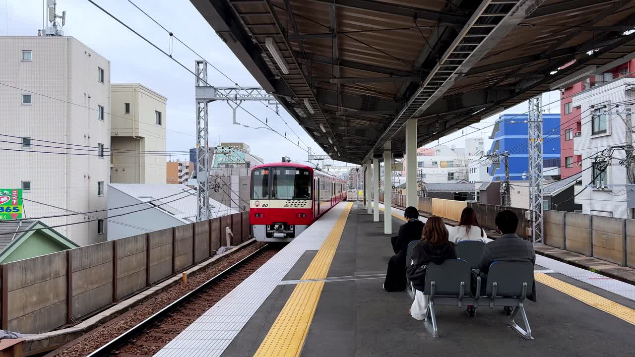 Passengers waiting on an outdoor train platform in Tokyo surrounded by city buildings on a cloudy day with train passing by