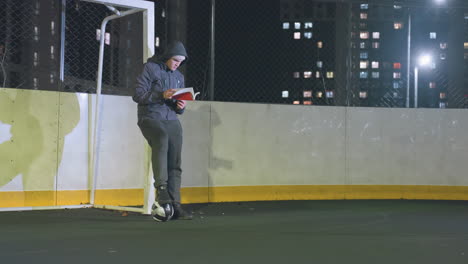 young boy in hoodie and beanie leaning against metallic goalpost reading book outdoors at night with soccer ball underfoot, scene illuminated by vibrant urban city lights