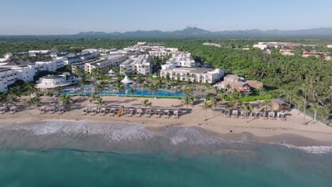 Vista-Aérea-Cinematográfica-Que-Muestra-Un-Hotel-Resort-En-Un-Paisaje-Pintoresco-En-La-Isla-De-La-República-Dominicana-Con-Playa-De-Arena---Olas-Del-Mar-Caribeño-Alcanzando-La-Costa-Al-Atardecer