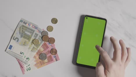 Disparo-De-Moneda-Aérea-Billetes-Y-Monedas-De-Euro-Junto-A-La-Persona-Que-Usa-Un-Teléfono-Móvil-Con-Pantalla-Verde