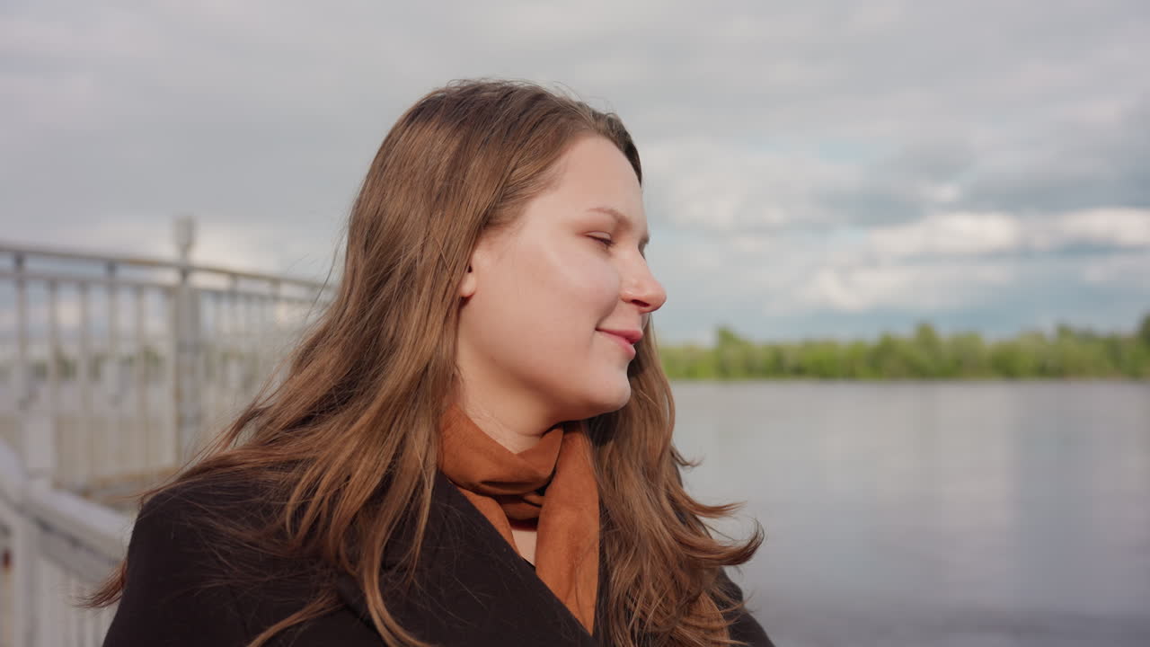 Scenic viewer gives sudden soft laugh while looking forward near calm river under gentle daylight, relaxed expression reflecting light joy as wind moves through hair creating natural serene mood