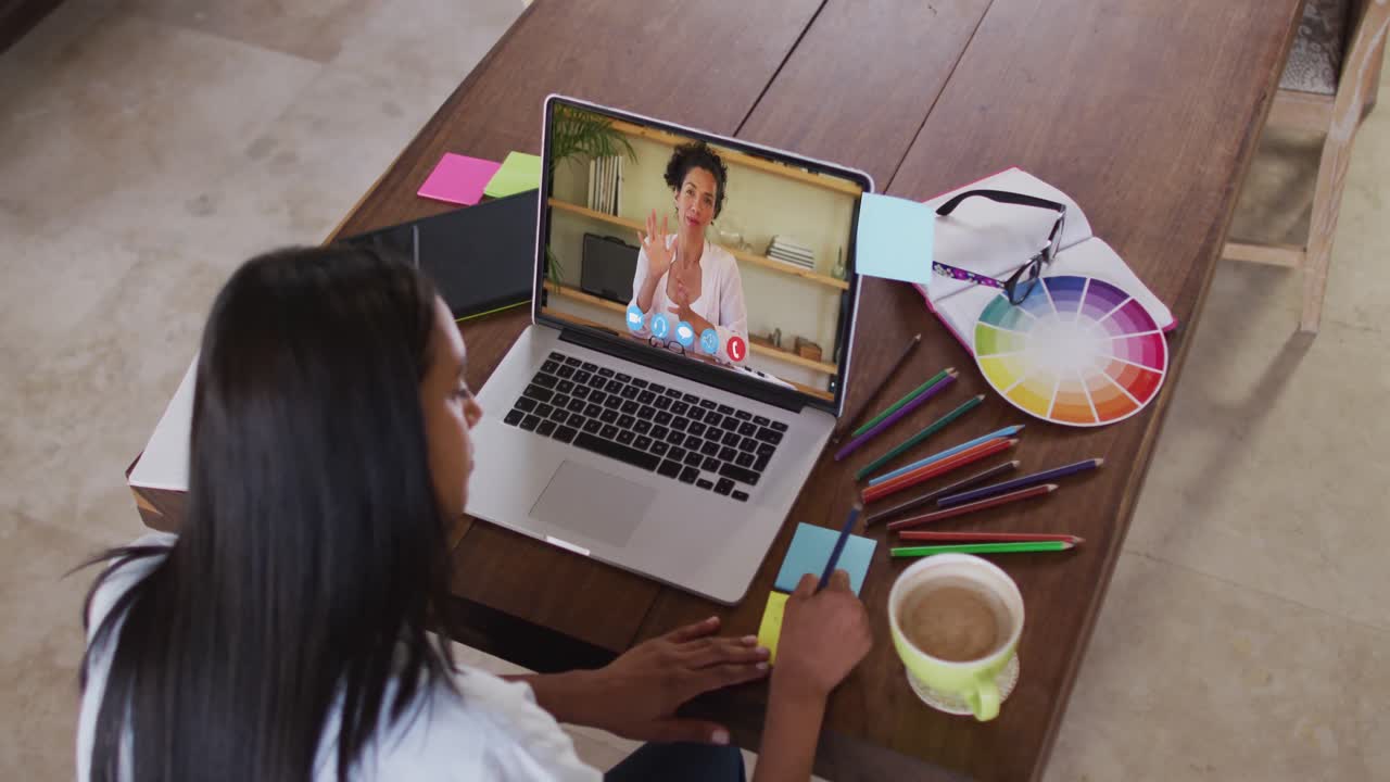 Mujer afroamericana escribiendo en notas adhesivas mientras hace una videollamada en su portátil en casa.