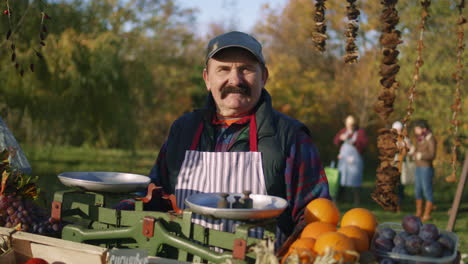 elderly farmer stands at the stall and smiles elderly farmer stands at the stall with fresh colorful fruits and vegetables looks at camera weekend shopping at local farmers market outside vegetarian organic and healthy food agriculture