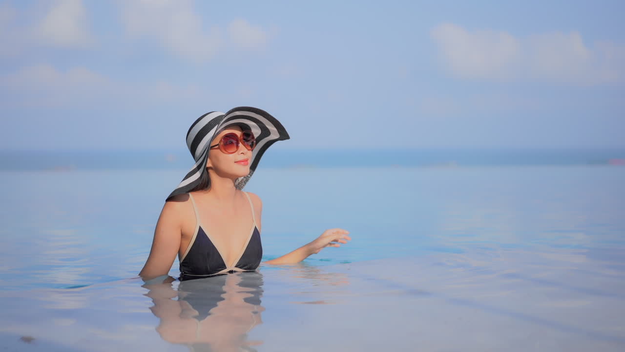 Static wide shot of a beautiful lady standing in an infinity pool wearing a beach hat and posing