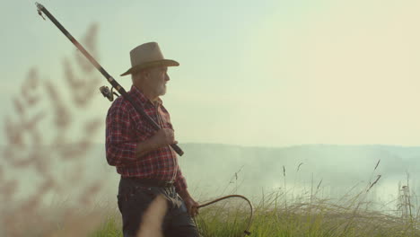 side view of old man in a hat standing on the shore of a lake with a fishing rod on a cloudy morning