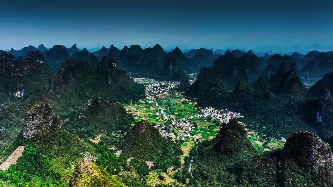 Green agricultural fields and village rooftops spread across a flat basin beneath steep dark mountains.