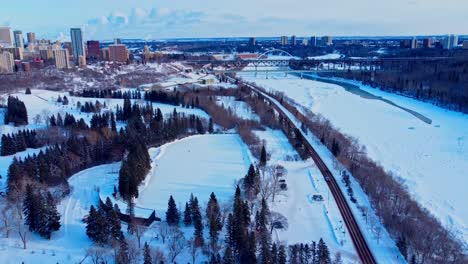 aerial flyover winter victoria park manmade quiet ice rink connected to an infinity skate track next to forest road next to snow covered icy north saskatchewan river edge of condos downtown city1-4