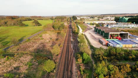 Tren-Acercándose-Y-Pasando-Bajo-Un-Disparo-De-Drone-En-Una-Vía-Con-Campo-A-Un-Lado-Y-Almacenes-Y-Polígono-Industrial-Al-Otro