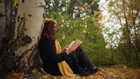 woman dressed in yellow beret and scarf sits against tree in autumn forest, thoughtfully flipping through book pages, golden leaves scattered around enhance peaceful, reflective atmosphere