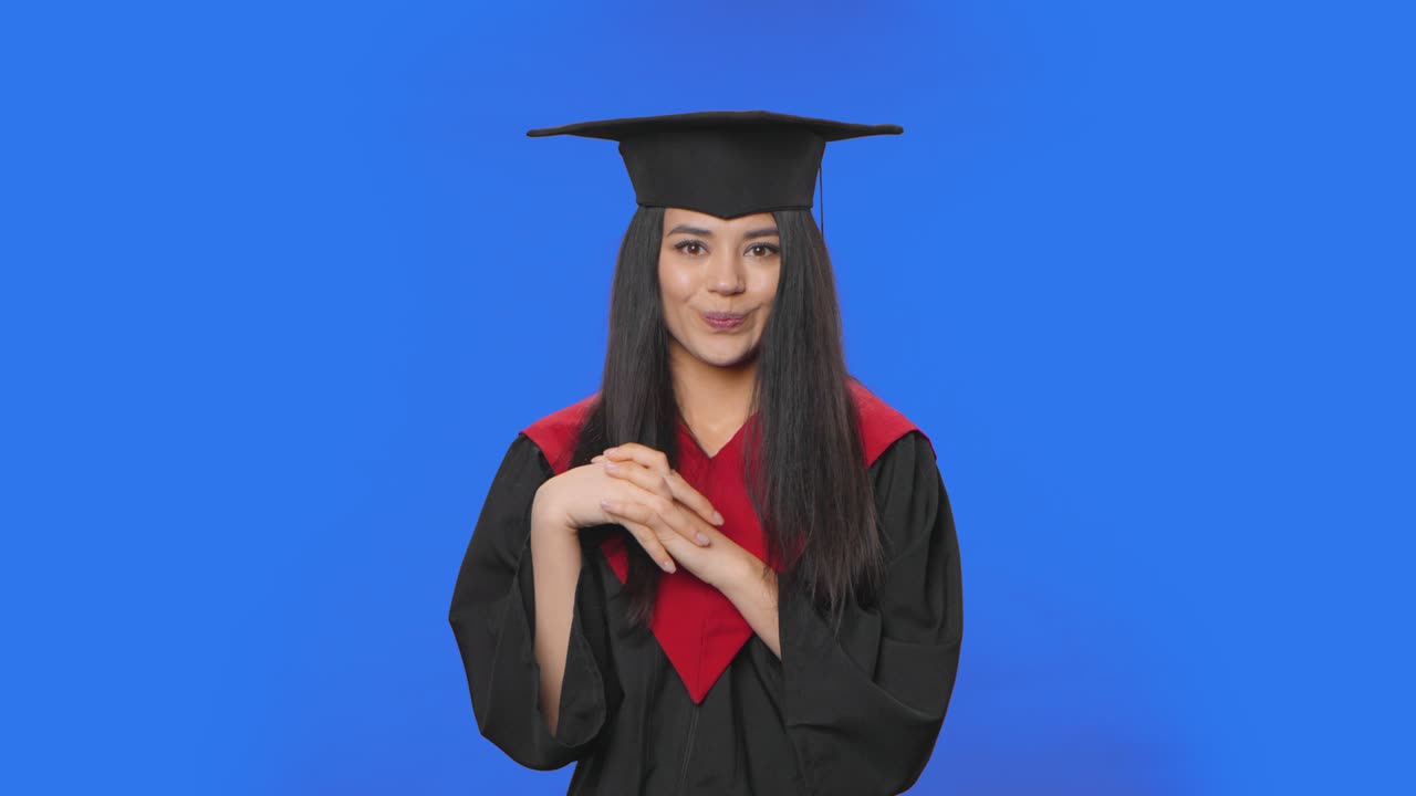 Portrait of confused female student in graduation costume saying oops, shrugging and biting lips. Young brunette posing in studio with blue screen background. Close up. Slow motion ready 59.94fps