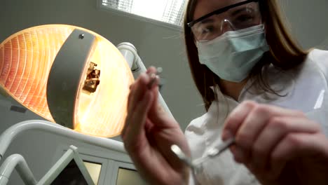 Young-Female-Dentist-in-Mask-Approaches-With-Tools,-Holding-a-Mouth-Mirror-and-Dentist's-Hook,-Dental-Lamp-Lights-Into-a-Patient