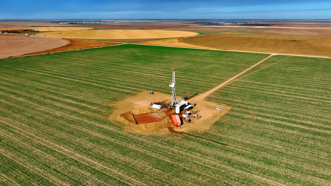 Big rectangular green field with drilling site in the middle. Drone footage around the oil production rig.