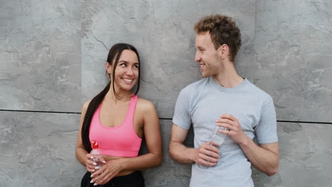 happy sportive couple leaning against a wall, holding water bottle and talking to each other, while taking a break during running session in the city