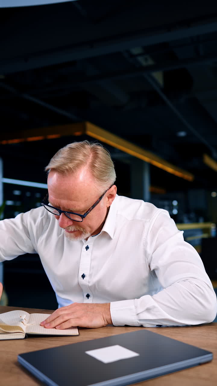 Mature Caucasian man in white shirt sits at desk. Businessman pulls the pages out of his notebook and throws the paper balls back. Vertical video.