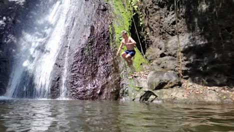 young man with blond hair jumping into water with tropical waterfall behind and making a splash