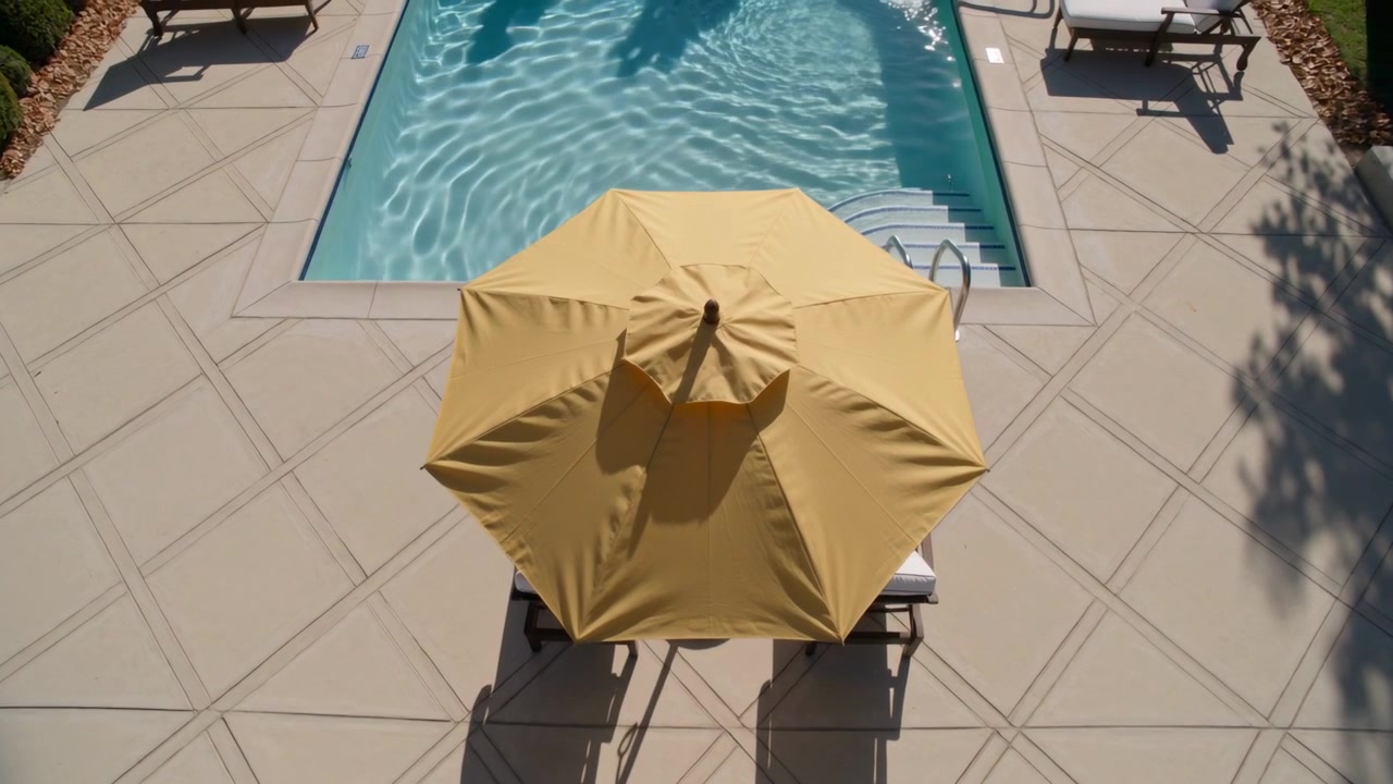 La luce del sole che si sposta proietta bagliori su un ombrellone giallo che ombreggia una sdraio nel patio della piscina, con increspature in movimento.