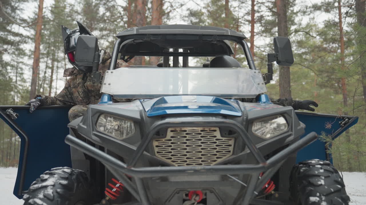 Due piloti ispezionano da vicino un ATV blu, aprendo lo sportello e controllando il verricello, con pneumatici robusti e paraurti visibili, mentre i compagni si preparano per un giro fuoristrada con i caschi indossati, sullo sfondo di pini innevati, effettuando controlli meccanici.