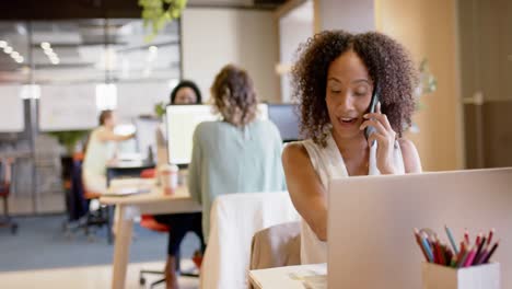 Biracial-Geschäftsfrau,-Die-Im-Büro-Auf-Dem-Smartphone-Spricht-Und-Einen-Laptop-Benutzt,-In-Zeitlupe