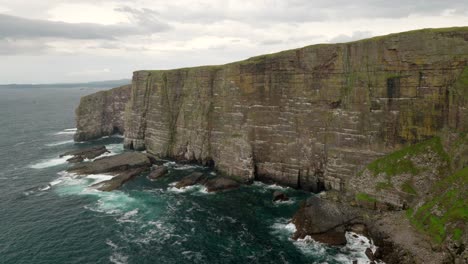 seabirds fly above a turquoise green ocean infront of a steep and dramatic cliff wall rising straight up out of the ocean as waves crash against its base