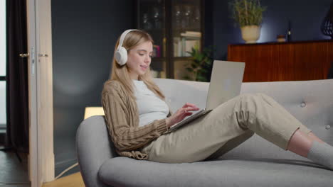 mujer joven feliz con auriculares inalámbricos charlando en la computadora portátil mientras está acostado en el sofá en casa 1