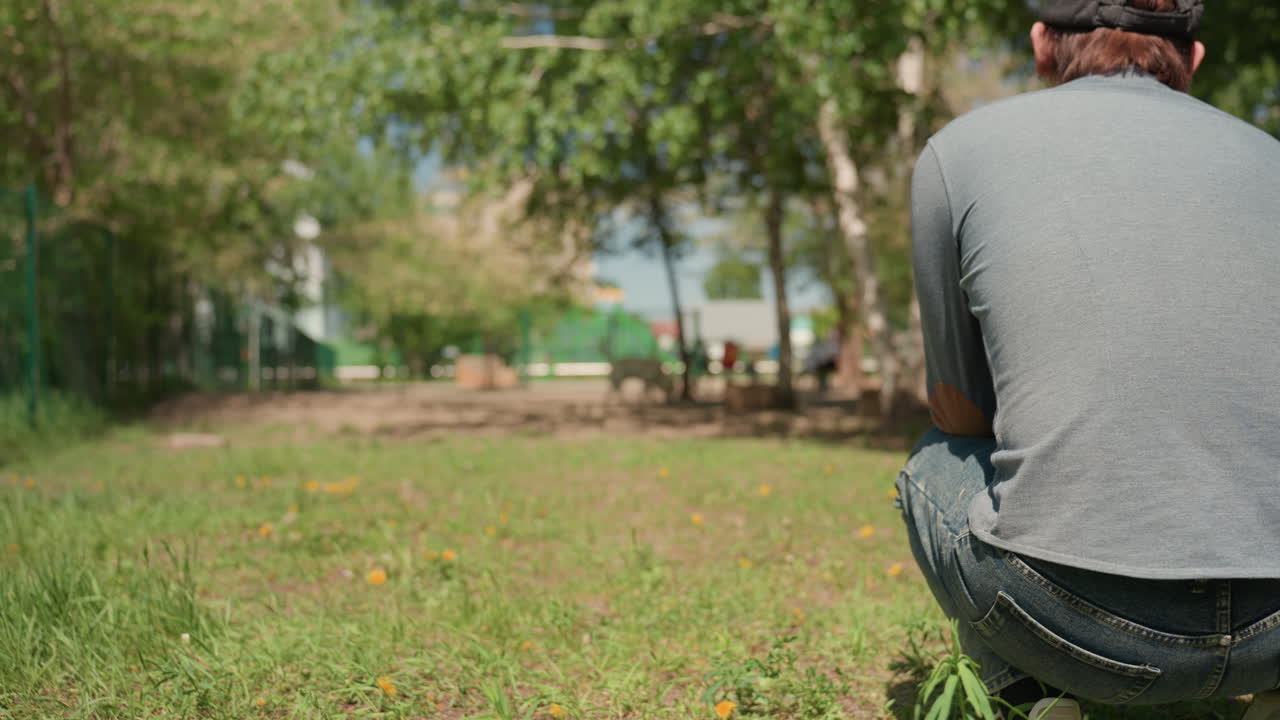조용한 관찰자가 기다립니다. 공원 경계에서 무릎을 꿇은 사람이 평화로운 멀리 떨어진 주변을 바라봅니다. 웅크린 형체가 주변 나무와 그늘 너머 멀리서 벌어지는 활동을 조용히 응시합니다.
