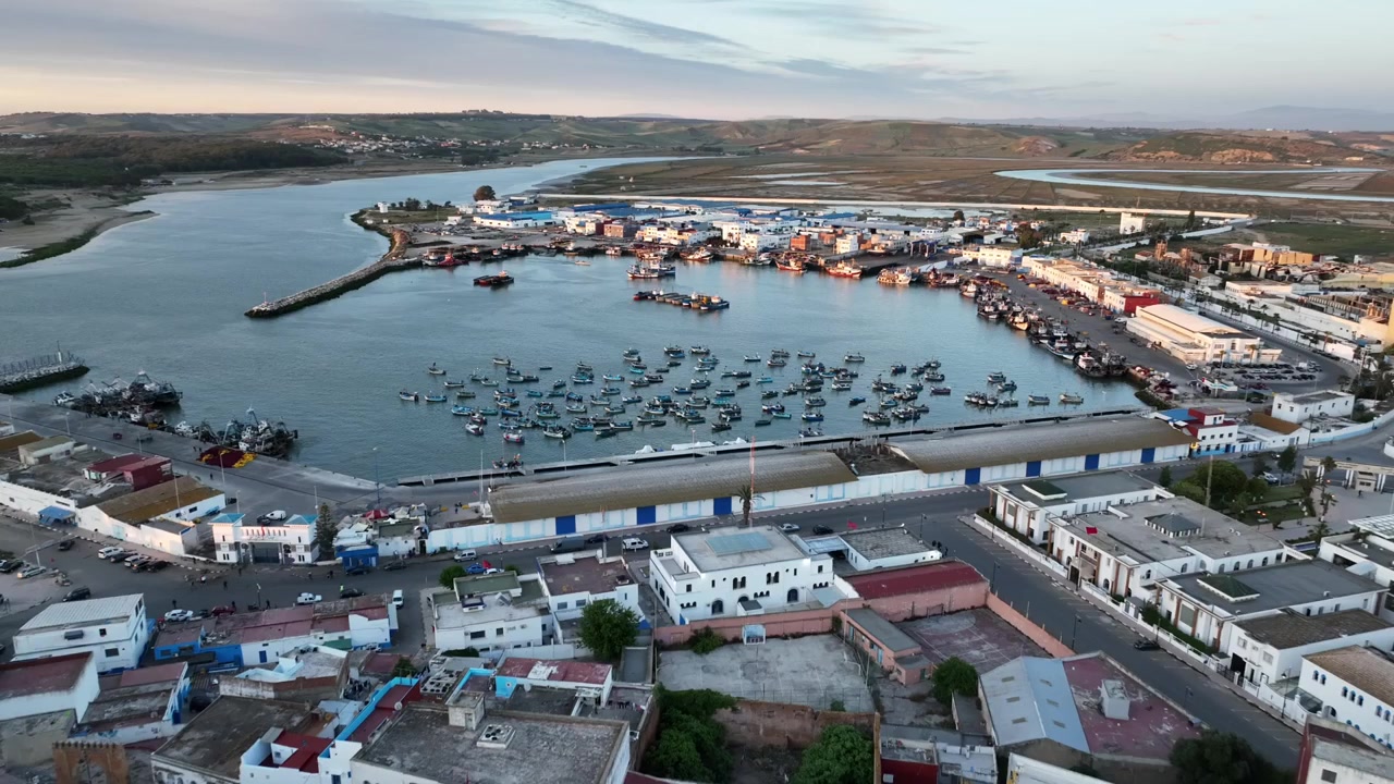 Vista aérea ampla do porto de pesca de Asilah em Marrocos, mostrando uma densa frota de pequenos barcos de pesca, instalações portuárias e a cidade costeira ao longo da costa atlântica.