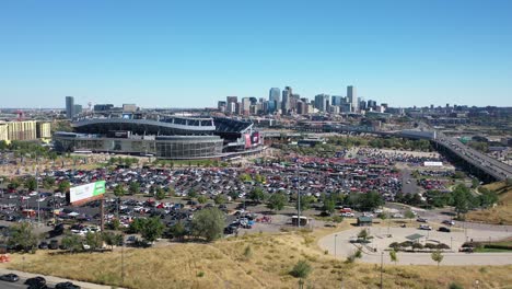 Vista-De-Seguimiento-Aéreo-Cinematográfico-Del-Campo-De-Empoderamiento-En-El-Estadio-De-Una-Milla-De-Altura-Con-Muchos-Autos-Estacionados-En-Espacios-De-Estacionamiento-Y-La-Autopista-De-La-Calle-Principal-Y-La-Vista-Del-Centro-De-La-Ciudad-De-Denver,-Colorado