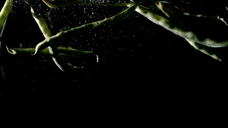 Green-bean-pods-dropped-into-water-on-black-background,-slow-motion-close-up