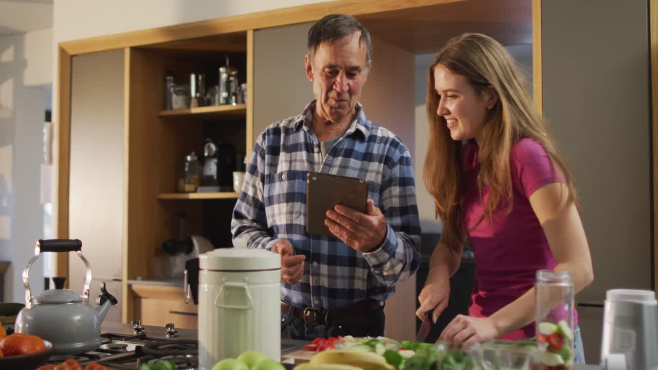padre mayor caucásico sonriente con su hija adolescente preparando comida y usando una tableta en la cocina