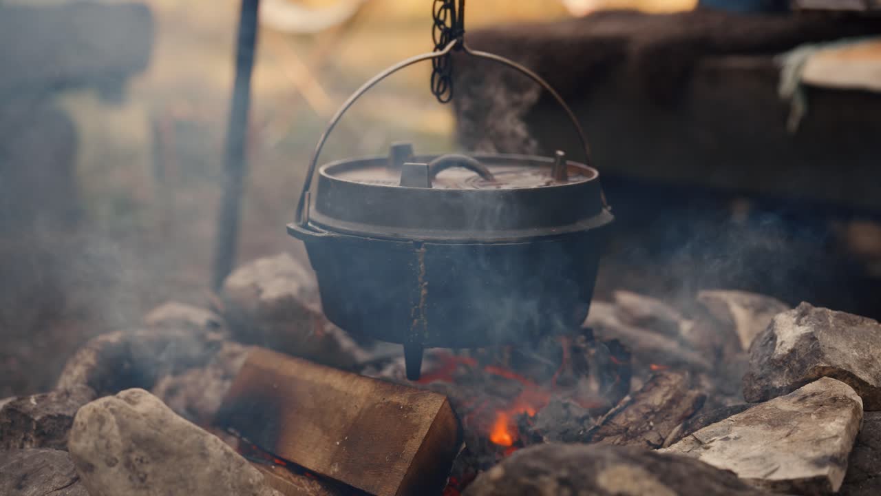 Rustic Dutch oven hanging above open campfire in forest with rising smoke and glowing wood logs