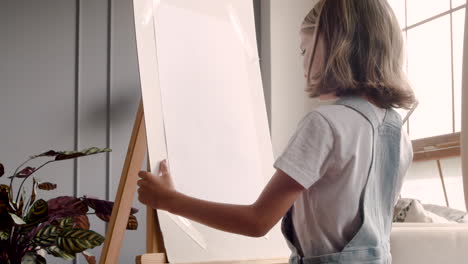 Rear-View-Of-Little-Blonde-Girl-Painting-On-The-Lectern-Next-To-The-Window-At-Home-2