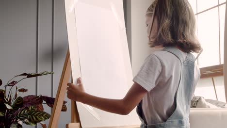 vista trasera de una niña rubia pintando en el atril al lado de la ventana en casa 2
