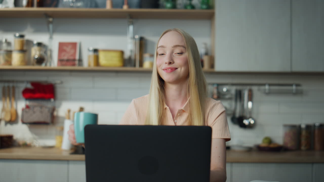 Pige chatter online på bærbar computer, sidder ved køkkenbordet og drikker kaffe i nærbillede