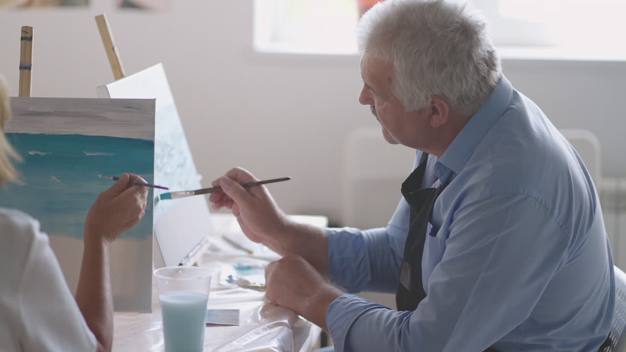 A female teacher shows a retired man how to draw a picture with paints and a brush at courses for the elderly. A senior man draws a picture to a group of pensioners
