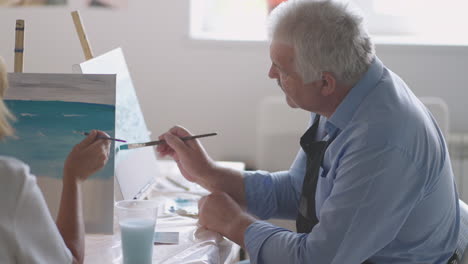 a female teacher shows a retired man how to draw a picture with paints and a brush at courses for the elderly. a senior man draws a picture to a group of pensioners