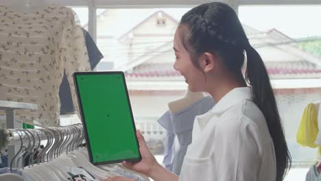 close-up de la mujer asiática dueña de una tienda de ropa sosteniendo y mirando el modelo de tableta de pantalla verde mientras comprueba las existencias en los rieles