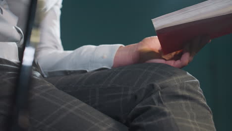 primer plano de una persona sin rostro con camisa blanca y pantalones grises sosteniendo un libro y un palo de clave. las manos descansan en el regazo con los dedos agarrando el palo de señal. la iluminación suave destaca las texturas de la ropa y los objetos