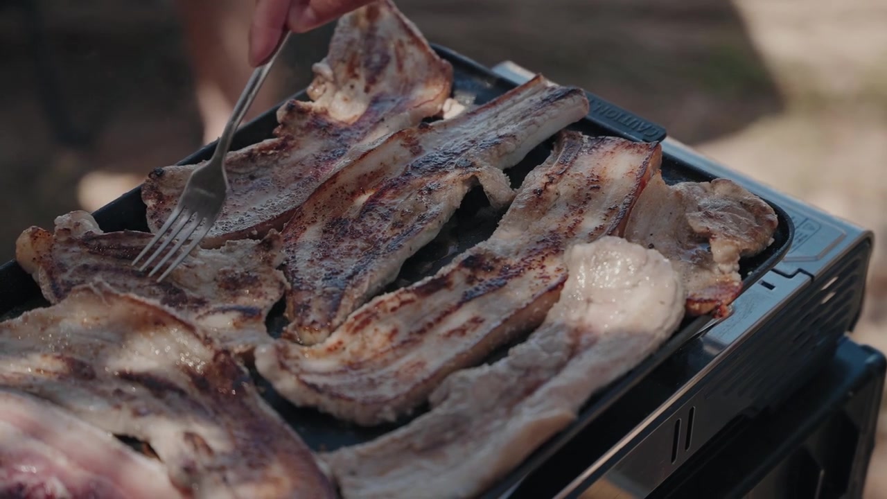 Forchetta che gira costolette di maiale grigliate e striscioline di pancetta che cuociono su una griglia barbecue nera da esterno