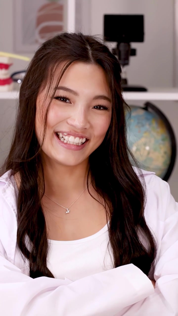 Confident teenage student in white lab coat smiles at camera in science classroom setting.