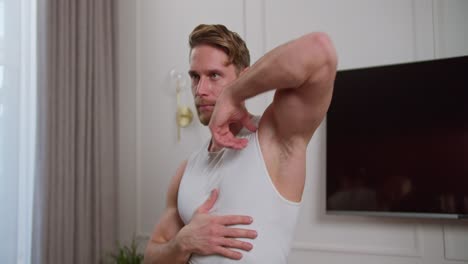 confident blond athletic man with stubble in a white t-shirt doing arm swings and stretching his shoulders before starting a workout at home in a modern apartment with a white wall