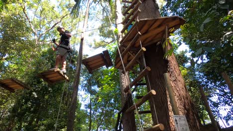 Ein-Glücklicher-Asiatischer-Mann-Tritt-über-Eine-Hoch-Hängende-Holzseilbrücke-In-Einem-Baumwipfel-Abenteuerpark