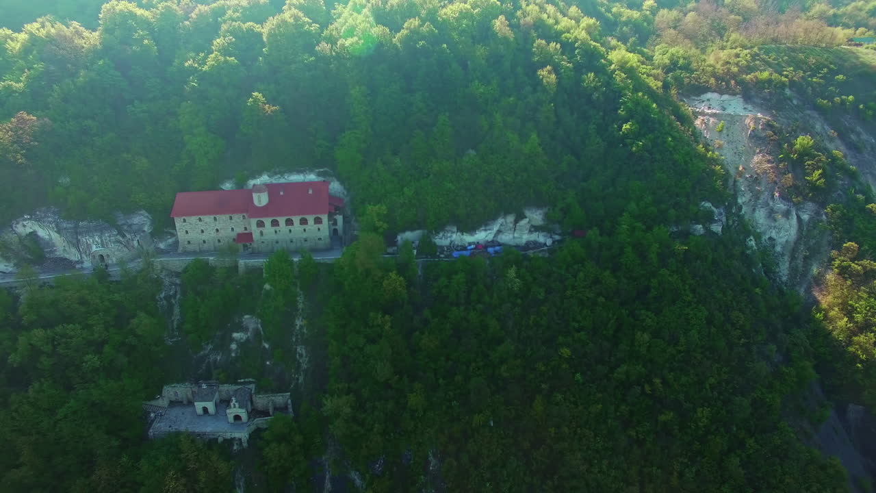 Orthodox rock monastery built on the sloping mount overgrown with pine trees. Christian church from aerial perspective on sunny day.