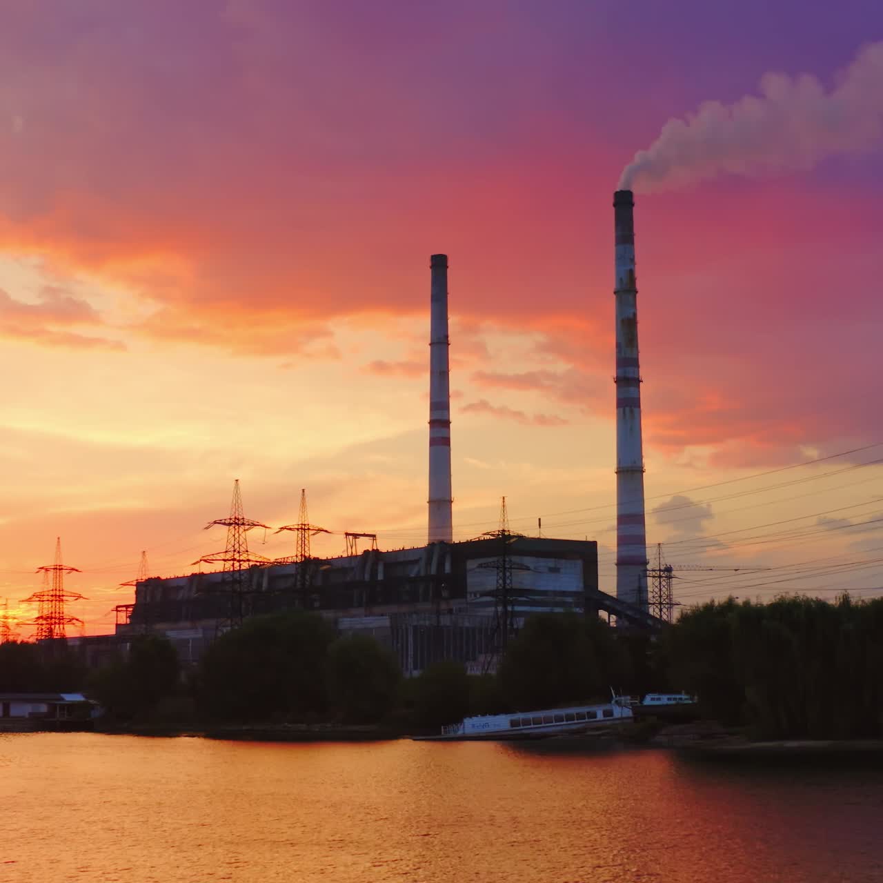 Manufacturing near the river. Industrial plant and high voltage electric towers among nature at sunset. View from the river. Aerial view.
