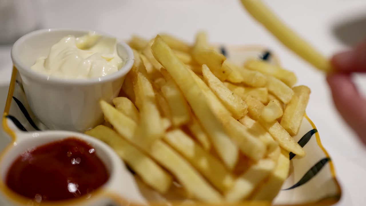 Uma mão mergulha batatas fritas em maionese e ketchup num prato listrado sob iluminação suave.